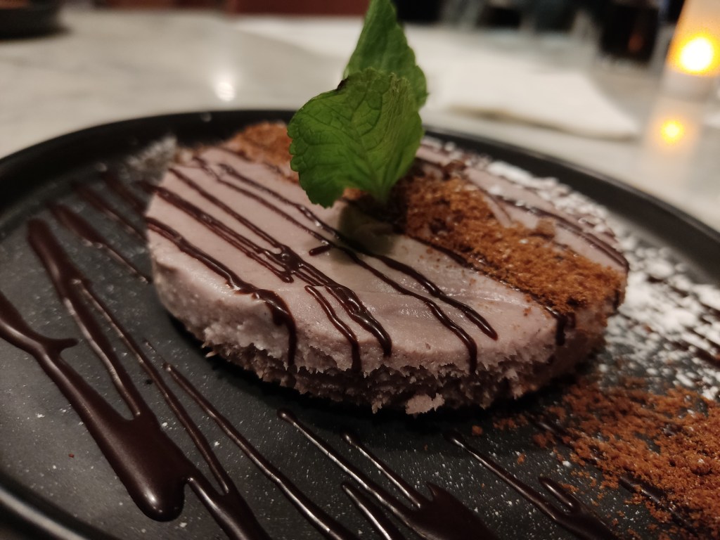 A round purple cheesecake with a very thin biscuit base, a line of cookie crumb across the top and chocolate sauce.
