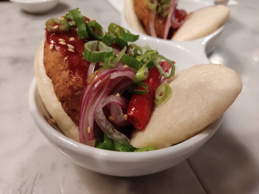 A bao bun with a fried chicken patty and some vegetables and sauce
