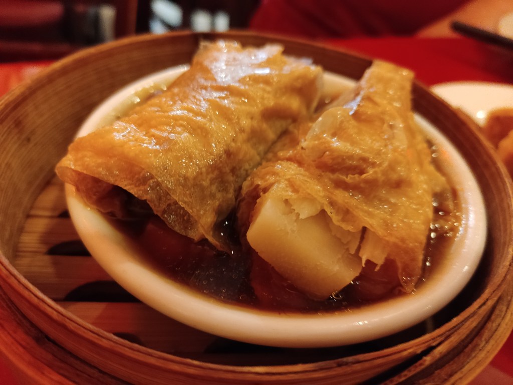 Two rolls covered in bean curd and sitting in a sauce, served in a steamer