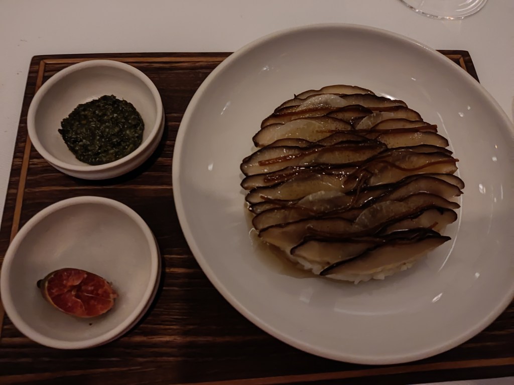 A large dish containing a circle of rice topped with slices of poached mushroom, with two small dishes on the side containing a fingerling lime and a dollop of pesto