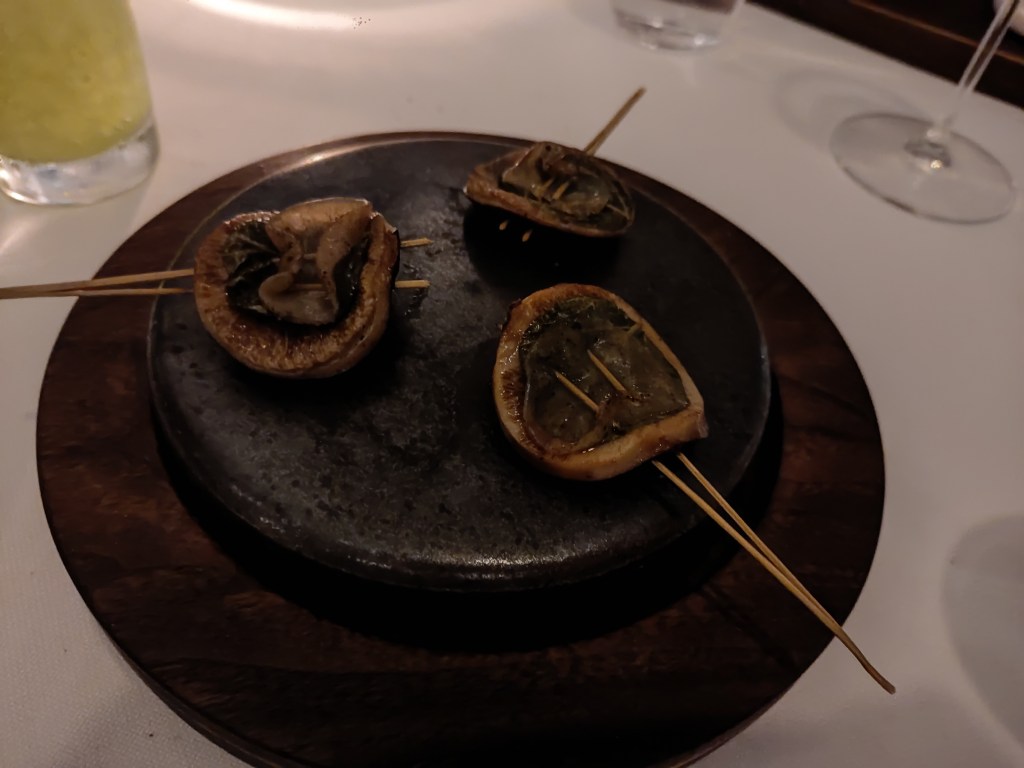 Three mushrooms on small wooden skewers, grilling on a hot stone in the centre of the table