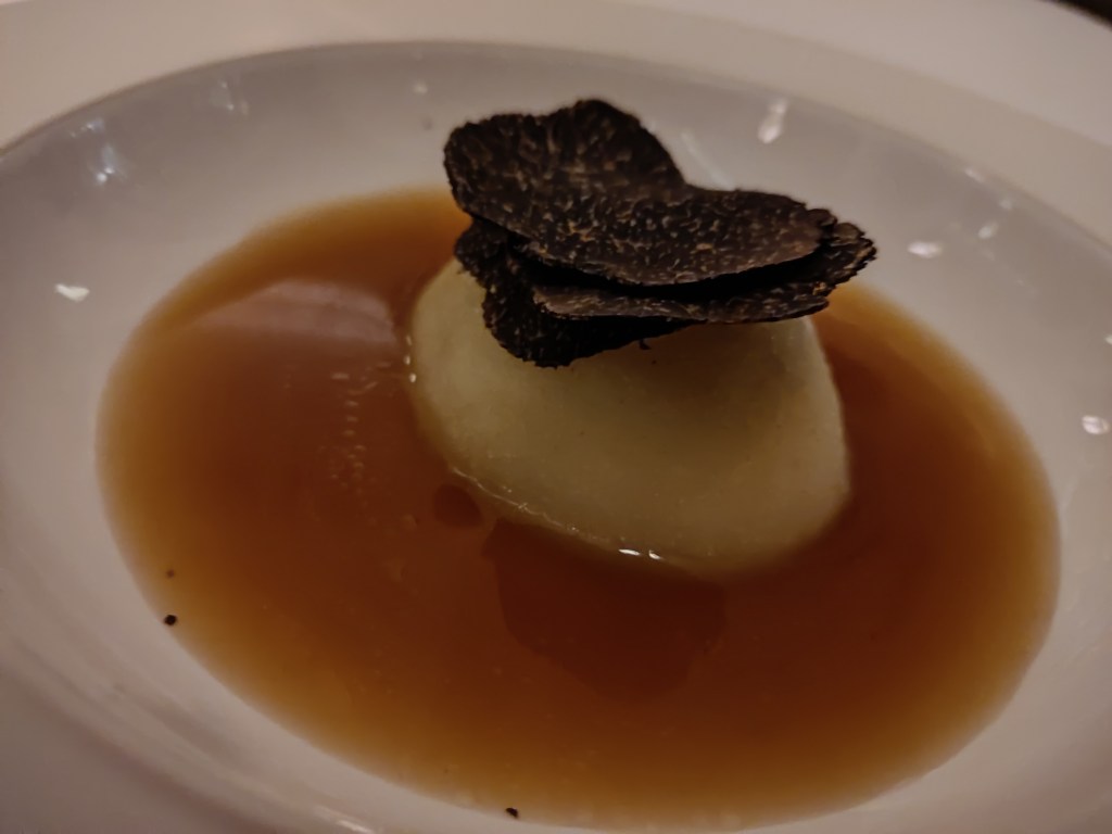 A volcano shaped potato, sitting in a broth and topped by large shavings of black truffle