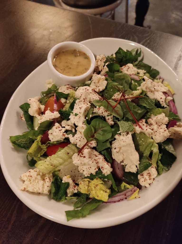 A large dish containing Greek salad including some creamy looking feta and a pot of dressing