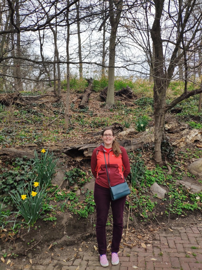 Ms HH smiling at the camera in Prospect Park, with some daffodils bursting into life