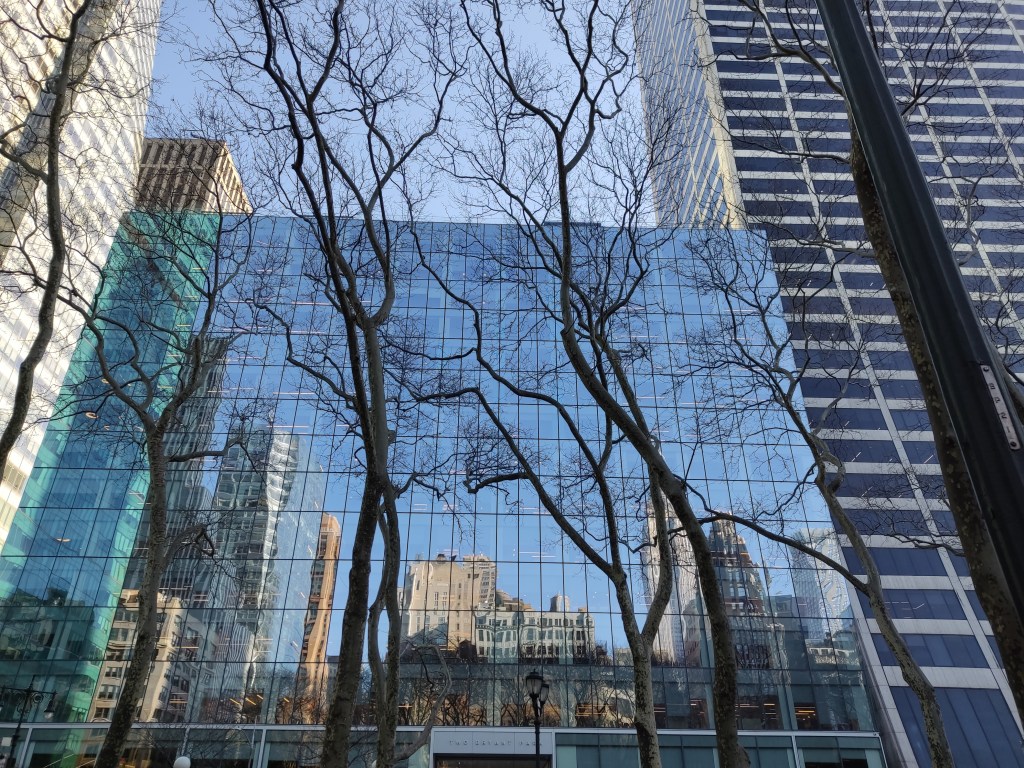 Skyscrapers reflected in a glass building facade, with bare trees in the foreground