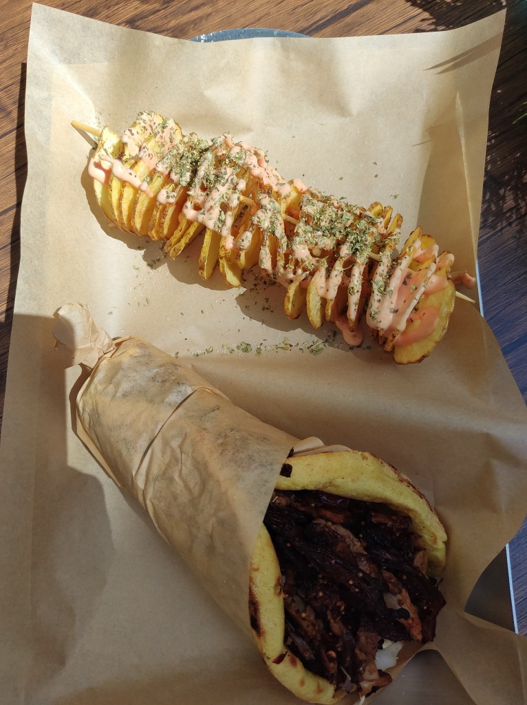 A wrap filled with meaty mushrooms, next to a spiralled potato on a long skewer, drizzled with a pink sauce and herbs