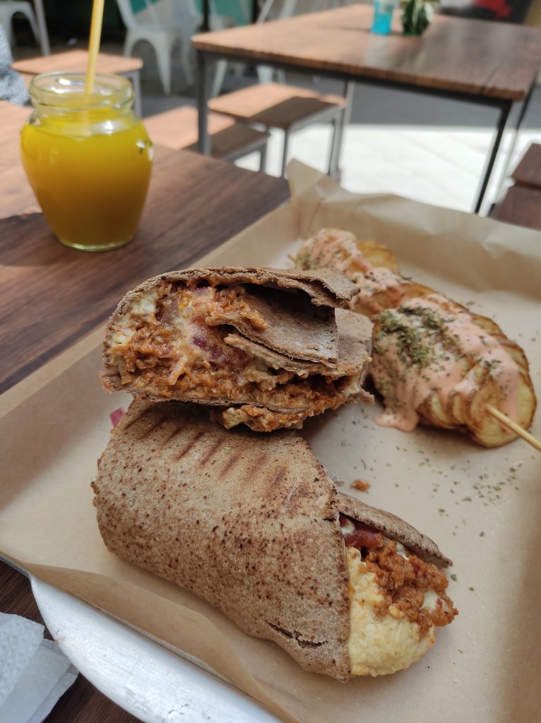 A wrap filled with sloppy looking mince and hummus. In the background there's a potato spiral on a stick, and a bright yellow drink