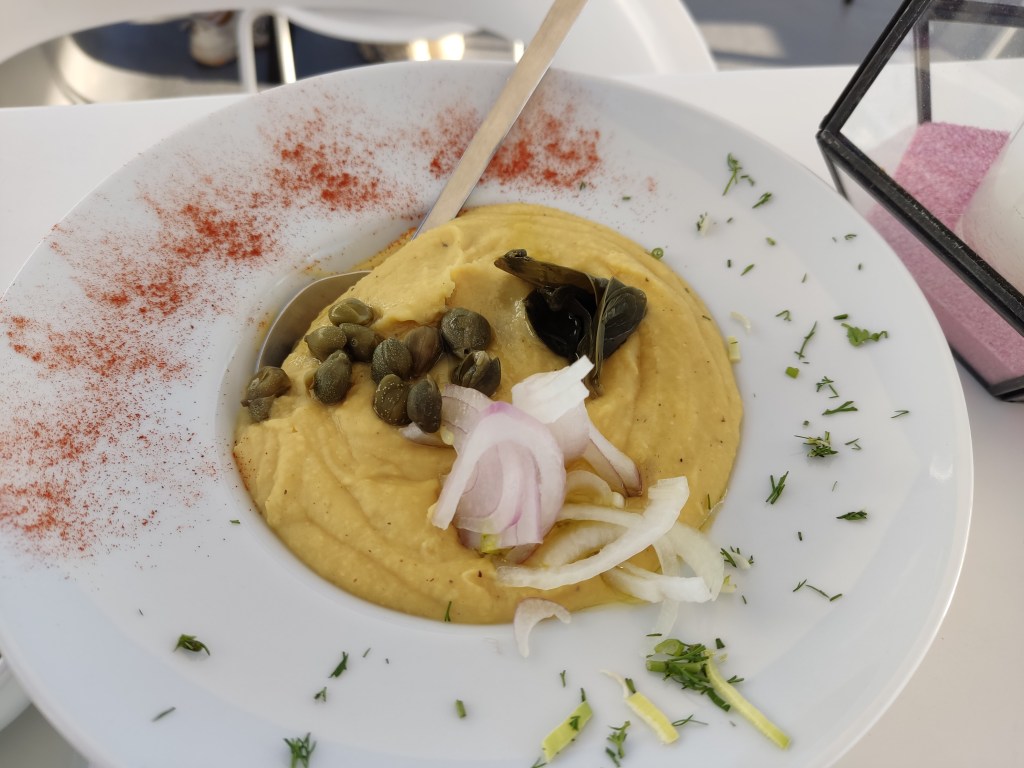 A dish with fava bean dip topped with capers, a caper leaf, and raw red onion