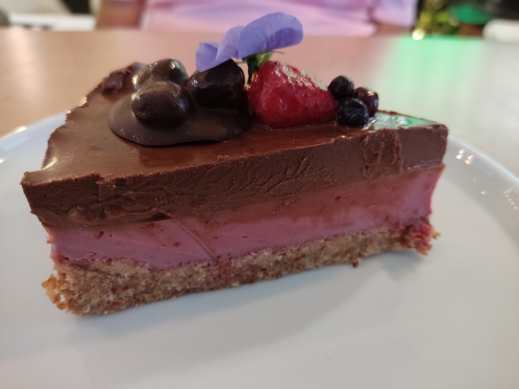 A cheesecake with a biscuit base, a raspberry layer, and a chocolate layer, topped with chocolate and fruit