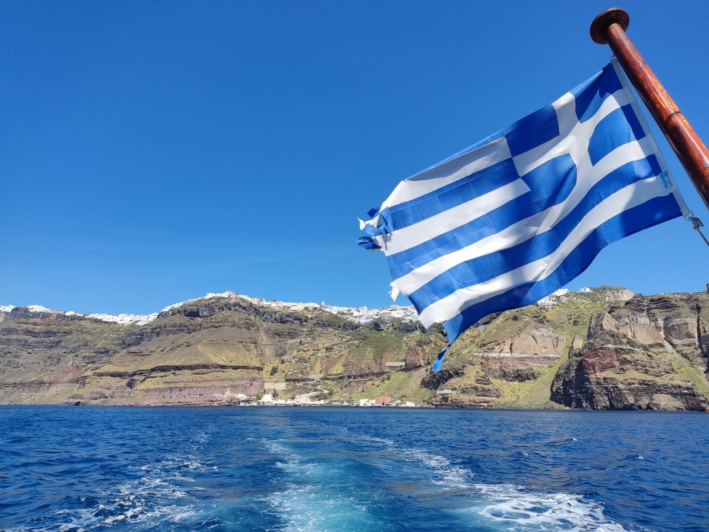 The Greek flag flying off the back of a boat as the sea stretches between the boat and Santorini