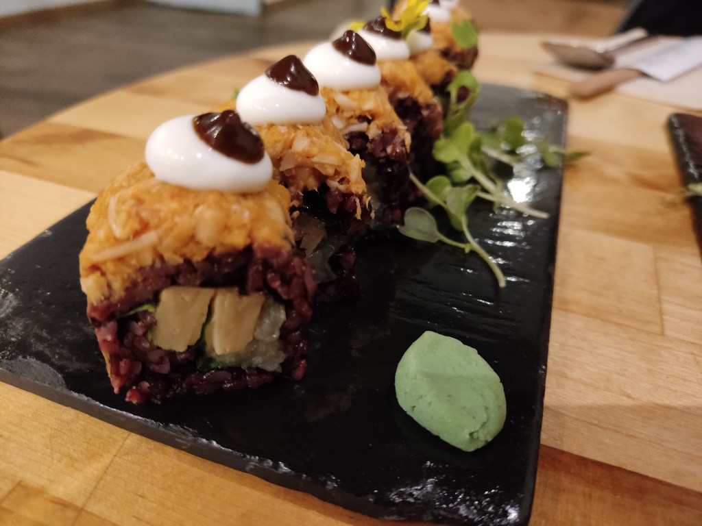 A slate with eight portions of sushi. The purple rice is on the outside, the filling is mock meat, and the topping is orange with a blob of white and then dark brown sauce on top