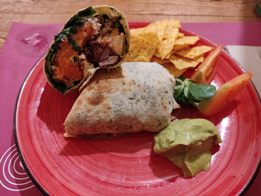 A burrito sliced in half filled with vegetables and grains, served with a dollop of guacamole, some tomatoes, and some crisps