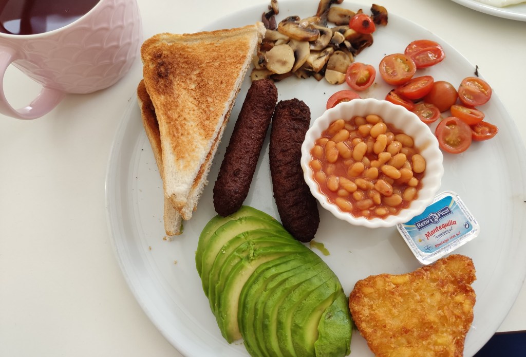 A big breakfast plate of toast, two sausages, a pile of fried mushrooms, a pile of halved cherry tomatoes, a pot of beans, a hash brown, and a sliced avocado