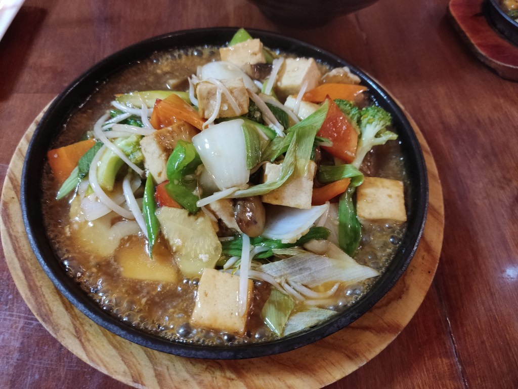 A big bubbling plate containing chunks of tofu along with onions, peppers, broccoli and other vegetables