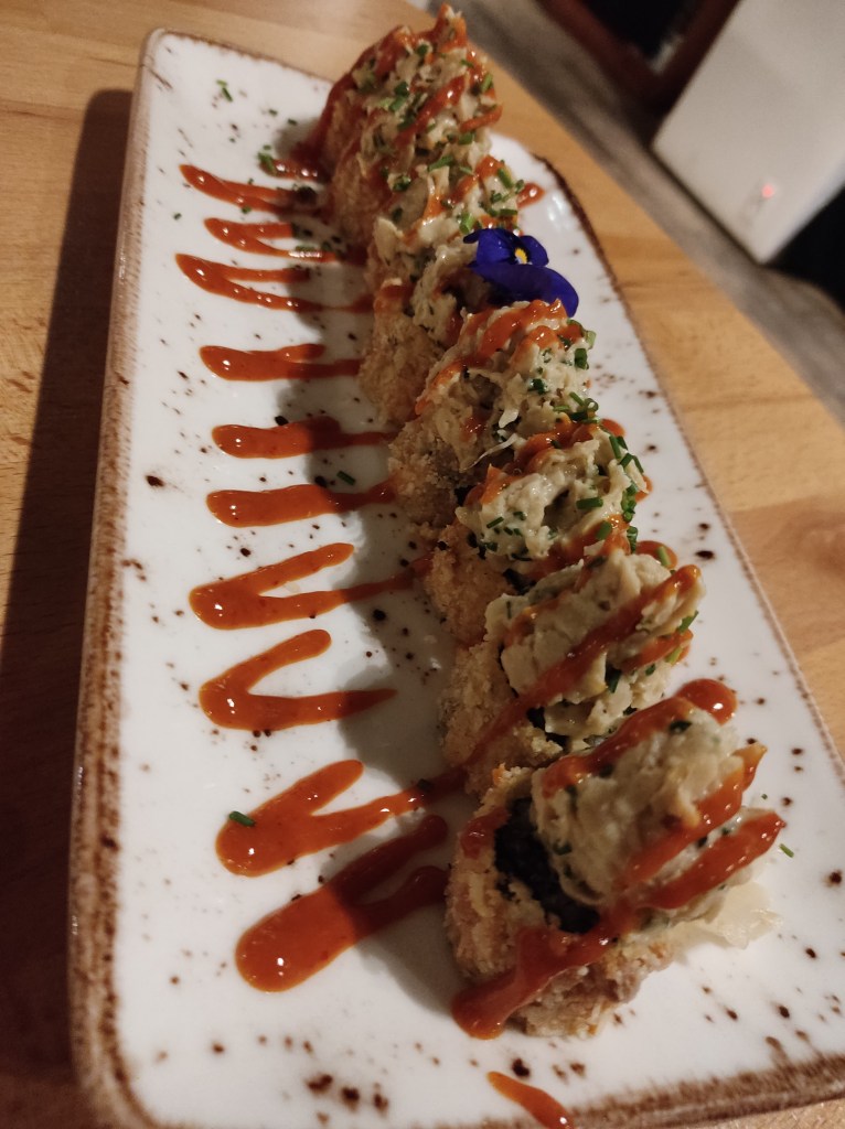 A plate with eight portions of tempura sushi, with a thick panko coating. They are piled with a beige cream and drizzled with a red sauce and herbs.