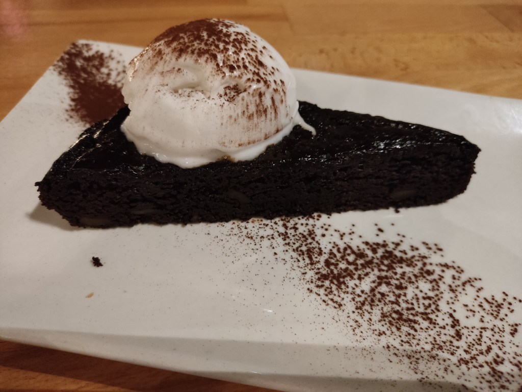 A slice of dark chocolate brownie topped with white ice cream and dusted with cocoa powder