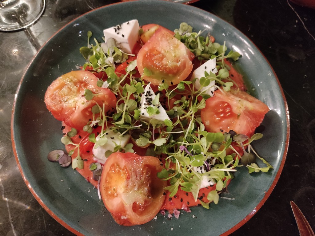 A plate with chunks of tomatoes and creamy white cheese on top of red sauce, scattered with micro greens and seeds