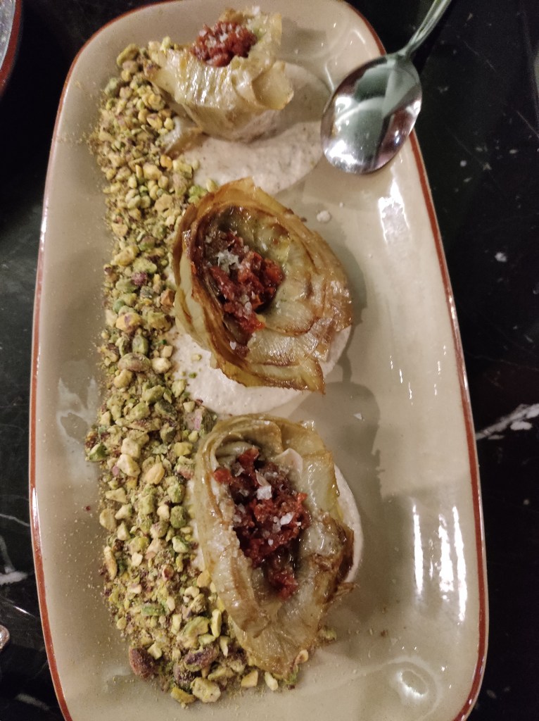 A long plate containing three artichoke halves filled with sundried tomatoes alongside green pistachio crumbs