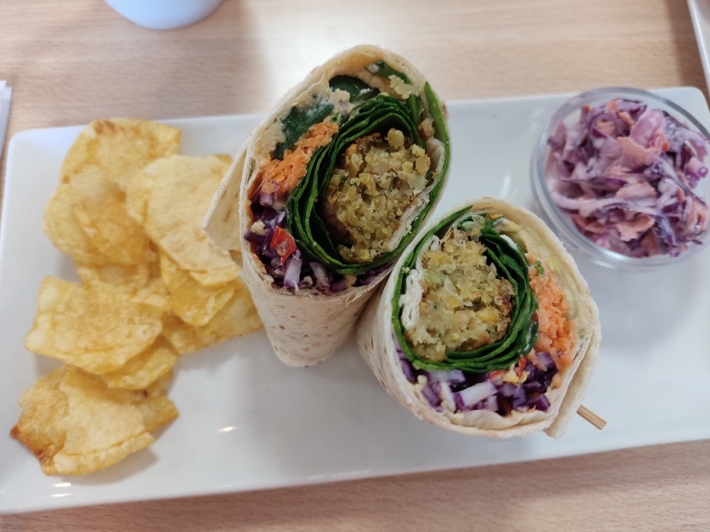 A wrap cut in half filled with falafel and salad, along with a handful of crisps and a pot of coleslaw
