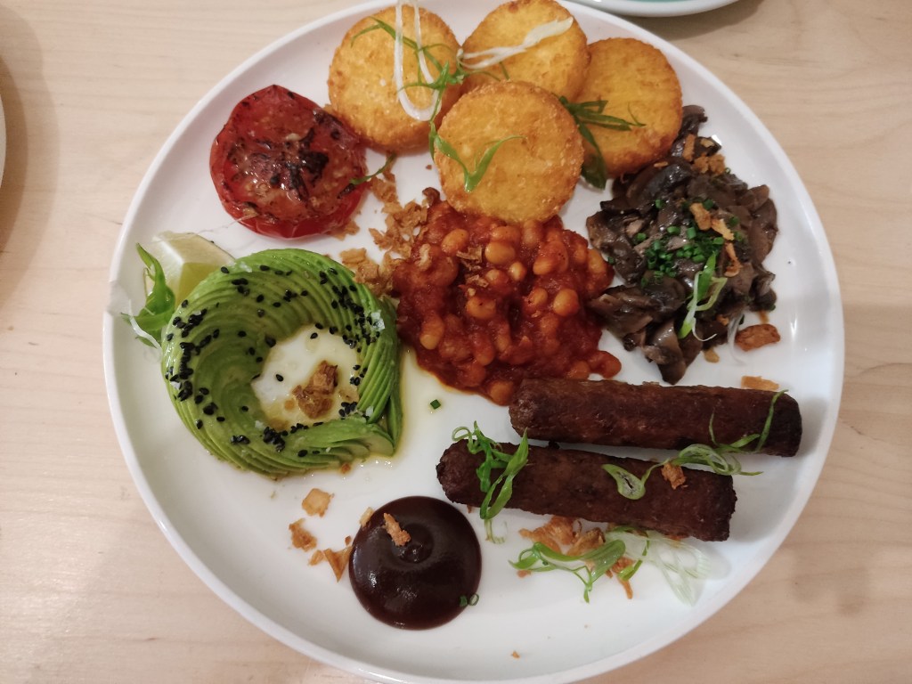 A big plate containing half a grilled tomato, four small hash browns, a pile of cooked mushrooms, two sausages, a dollop of brown sauce, an artful arrangement of sliced avocado with black sesame and lime, and a big helping of baked beans, with microgreens scattered over the plate 