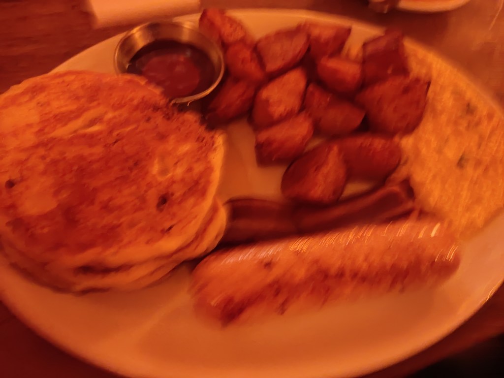 A stack of pancakes, a small pot of beans, some well roasted potato chunks, a small wet pile of tofu scramble, a small, crispy bacon rasher and a big juicy sausage