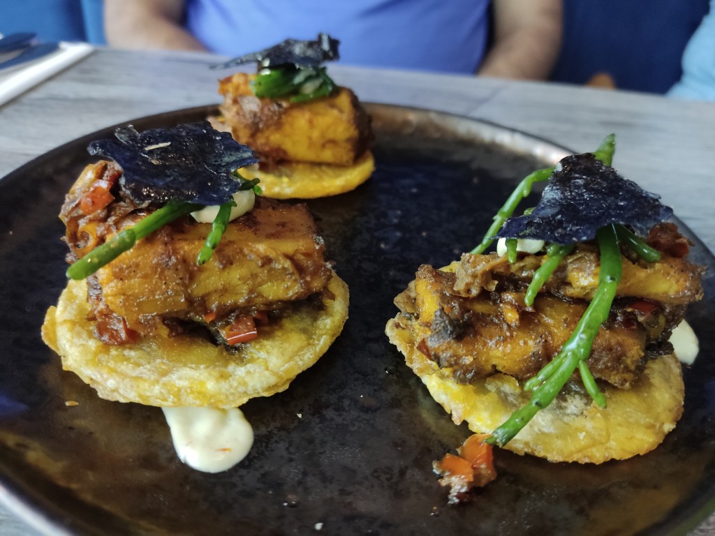 Three tostones on a plate topped with tofu fish and some greenery and seasonings