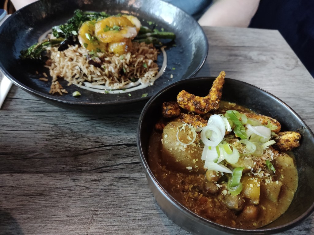 A plate of rice, broccoli and plantain in the background, with a pot of curry sauce, potatoes, and vegan prawns in the foreground