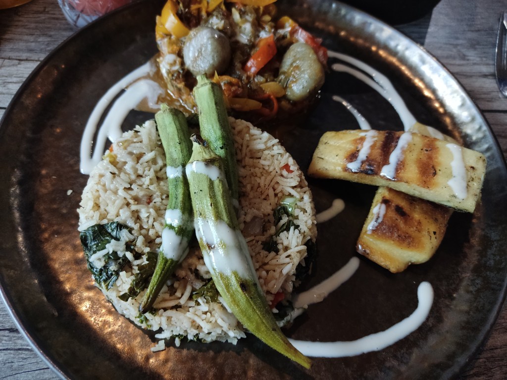 A mound of rice topped with okra with two big pieces of tofu fish on the side and a mound of peppers and dumplings in the background