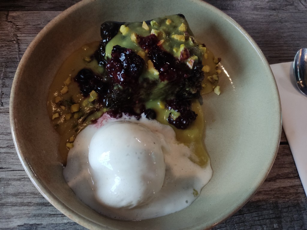 A bowl with a big scoop of ice cream, a puddle of yellow custard, and a square of dark ginger cake topped with raspberries and pistachio chunks