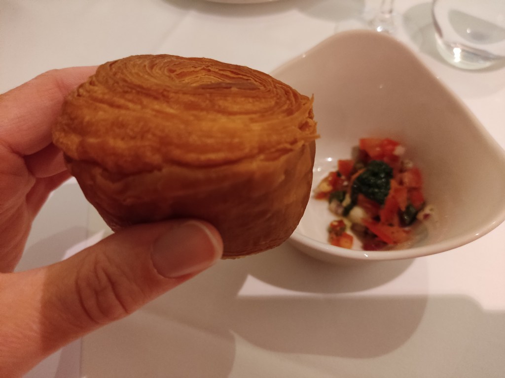 A beautifully layered bread or pastry in the foreground with a small dish containing tomato and seasoning in the background