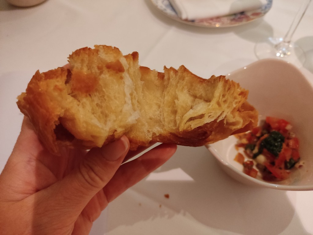 A bread or pastry in the foreground which has been opened up to reveal the layers, with a small dish containing tomato and seasoning in the background