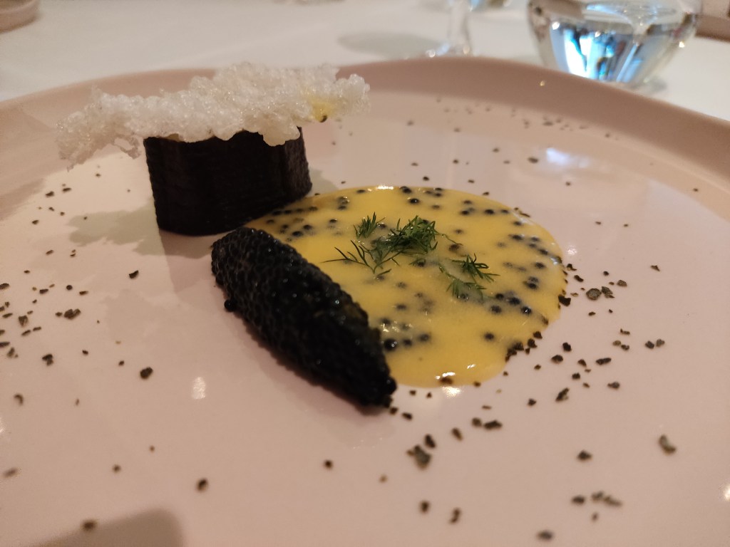 A quenelle of black seaweed caviar sitting alongside a buttery yellow sauce with black seeds and dill, alongside a seaweed wrapped oval, topped with a white textured cracker