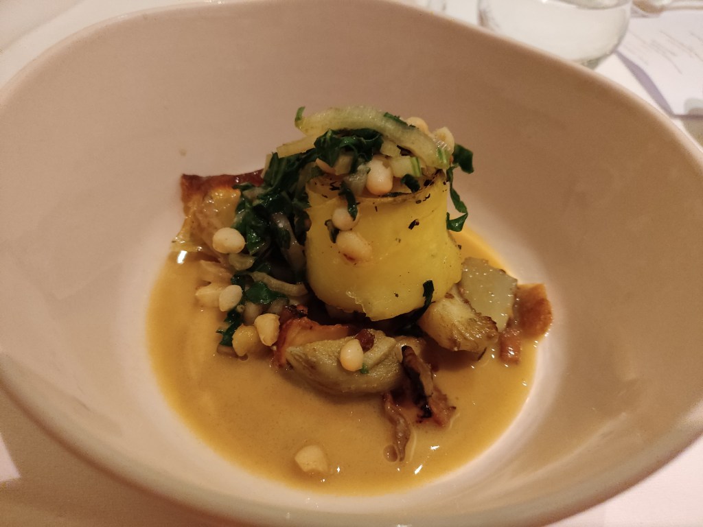 A long strip of potato rolled into a rose shape, topped with beans and greens, sitting atop a yellowish sauce with mushrooms and more beans