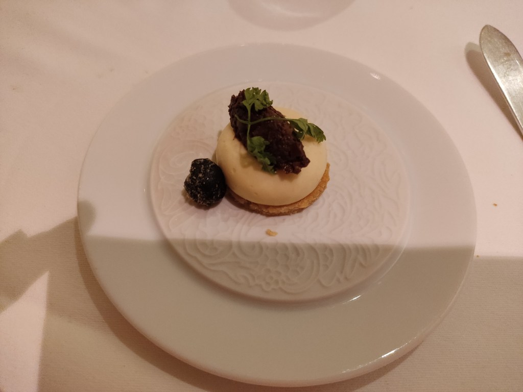 A small biscuit topped with smooth cream cheese and tapenade with two leaves of herb, and a caramelised olive on the side