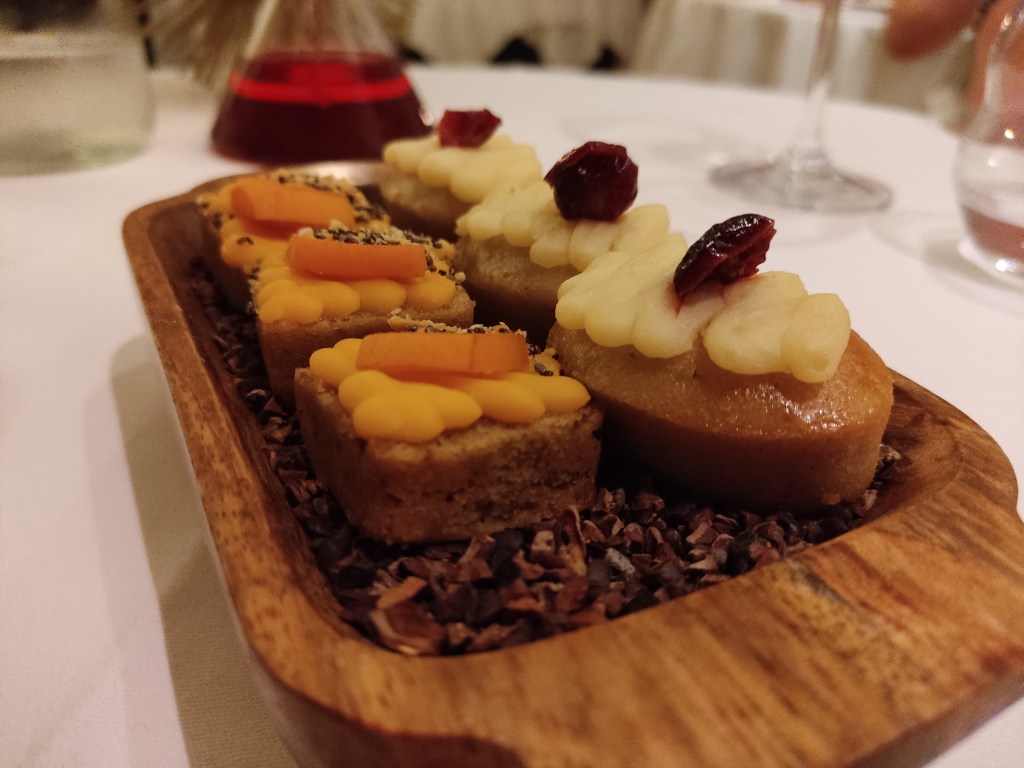 A tray containing three small cakes topped with yellow cream and apricot, and three small cakes with paler cream and a red fruit