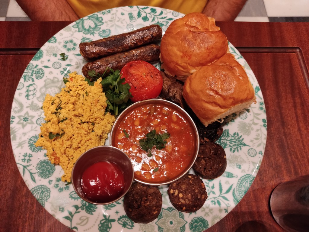 A big plate containing two sausages, a large helping of yellow tofu scramble, a pot of ketchup, a pot of baked beans, some rounds of black pudding, two bread buns, a tomato, some coriander leaves and a small glimpse of mushroom