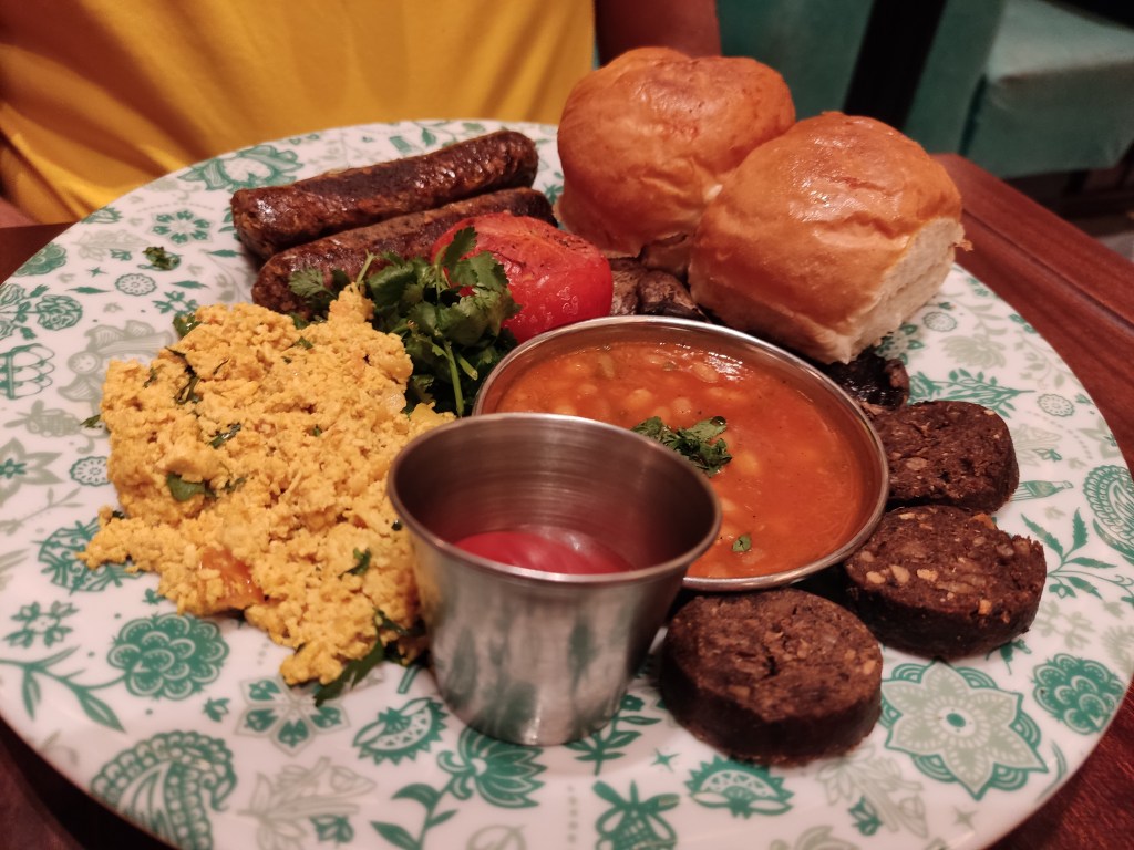 A big plate containing two sausages, a large helping of yellow tofu scramble, a pot of ketchup, a pot of baked beans, some rounds of black pudding, two bread buns, a tomato, some coriander leaves and a small glimpse of mushroom