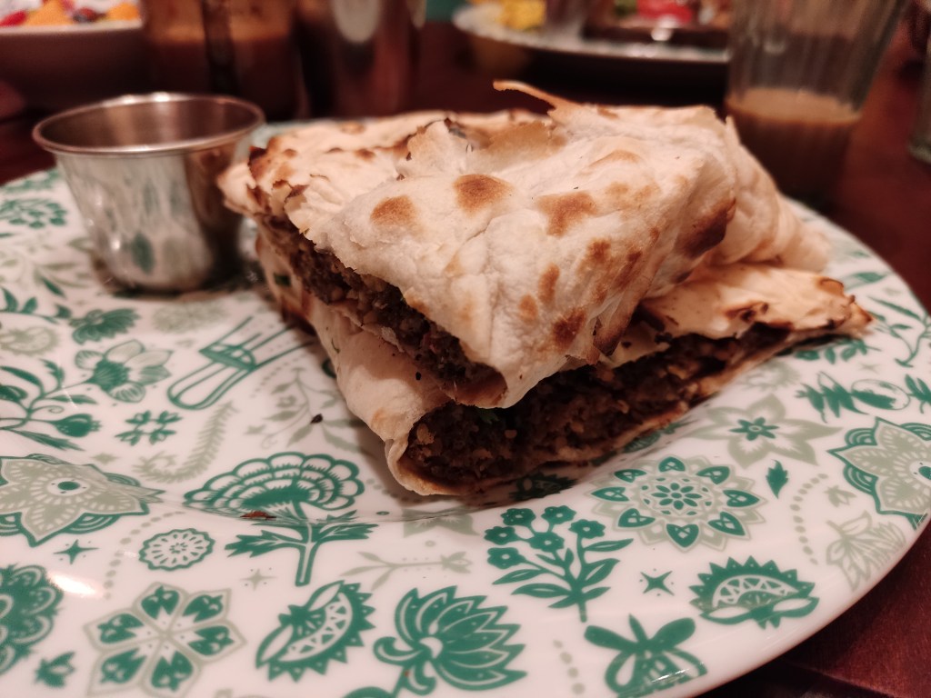 A plate with a pot of relish, and two pieces of a sandwich with naan folded around sausages