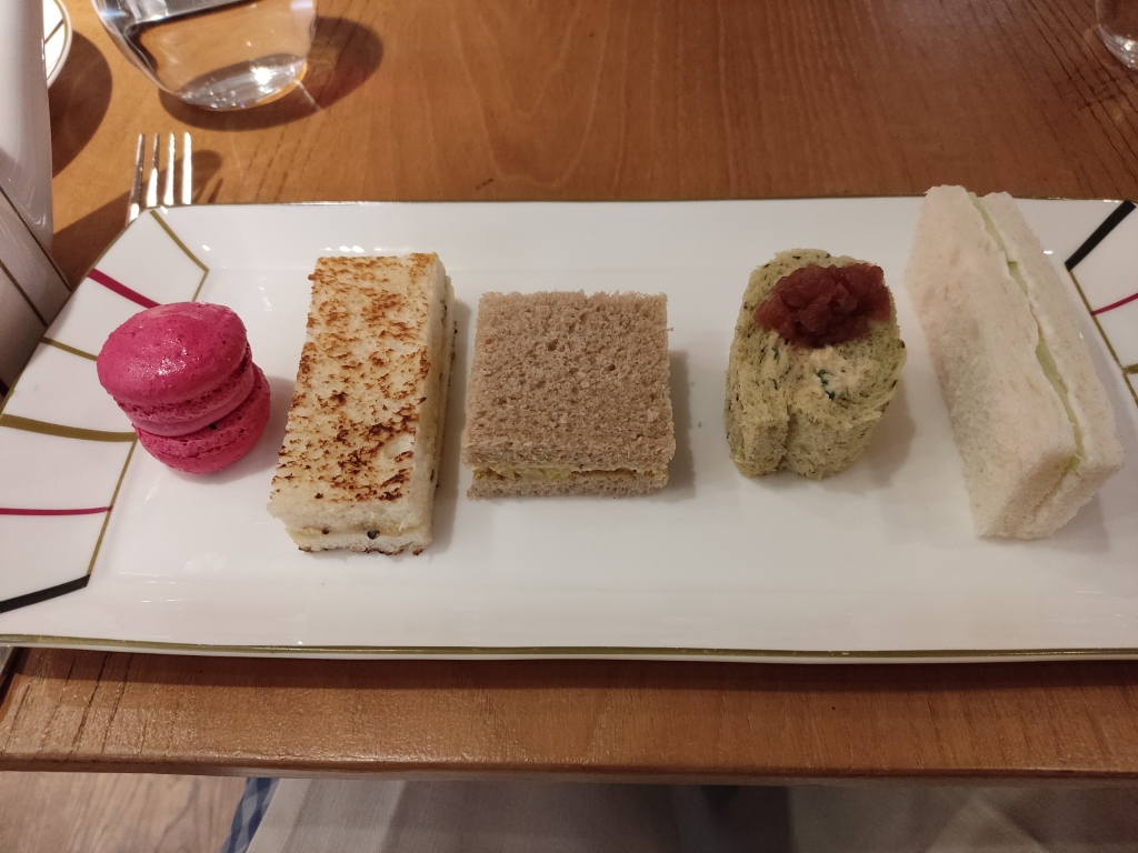 A plate containing a bright pink macaron, a toasted finger sandwich, a square sandwich, a rolled hummus sandwich topped with a dollop of chutney, and a white finger sandwich