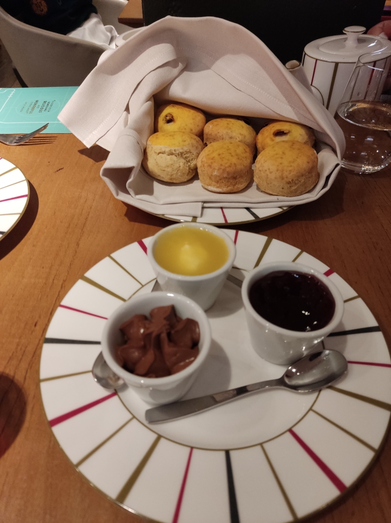 Six scones on a plate in the background, with three pots on a plate in the foreground, containing chocolate ganache, a bright yellow split cream and a red jam