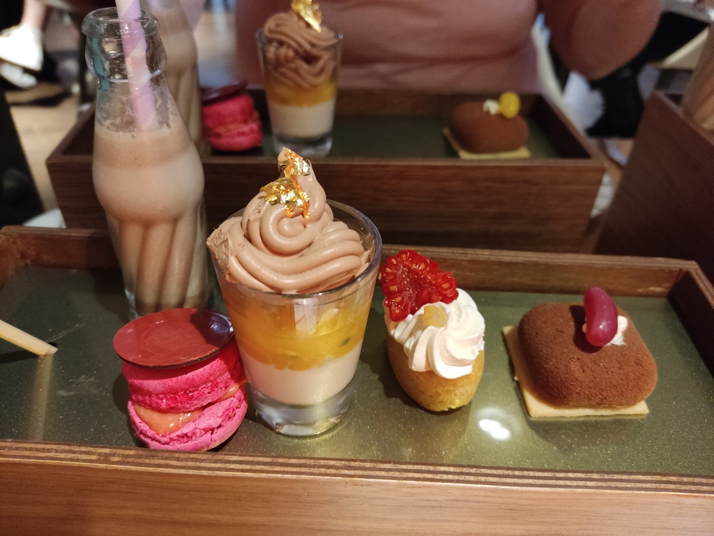 An assortment of sweets: a small glass bottle of chocolate milk with a straw, a bright pink macaron topped with a red disc, a small glass with three creamy layers and topped with gold leaf, a small oval sponge cake topped with cream and a raspberry, and a square chocolate sweet topped with a jelly bean
