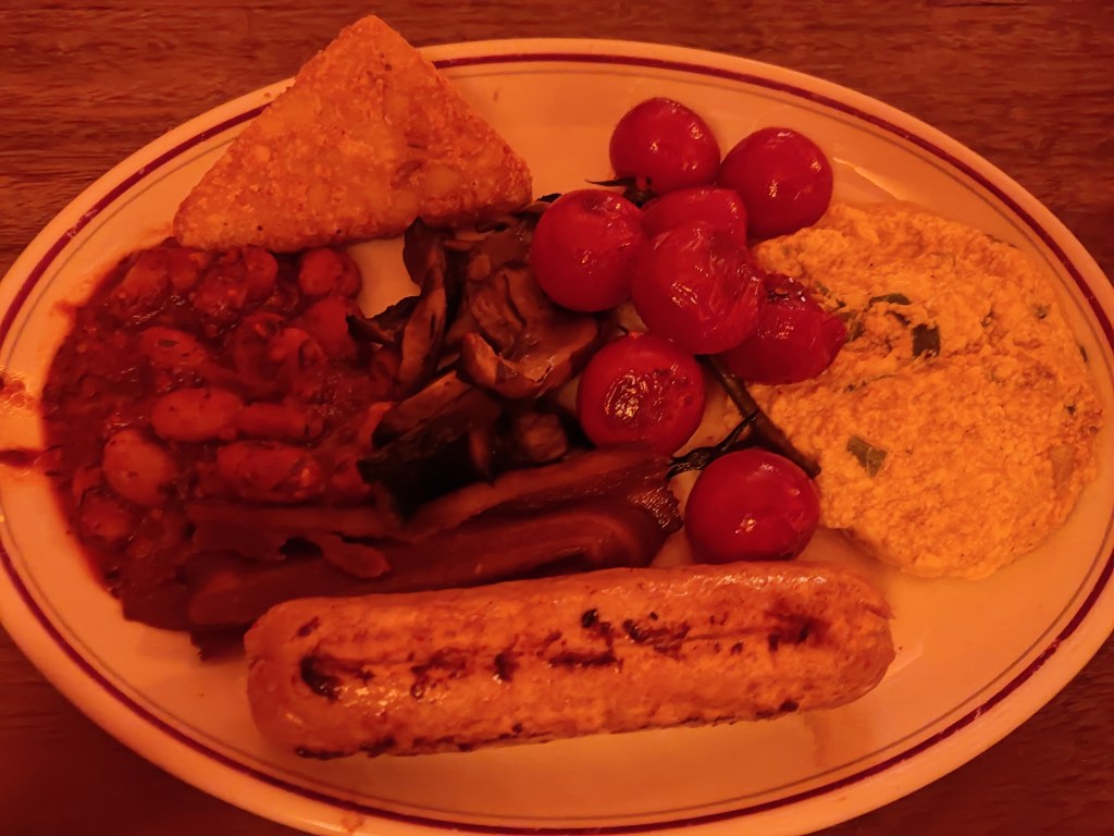 A plate containing a hash brown, pile of fried mushrooms, vine of roasted cherry tomatoes, a big wet pile of tofu scramble, a big juicy sausage, a small crispy bacon rasher, and a pile of beans
