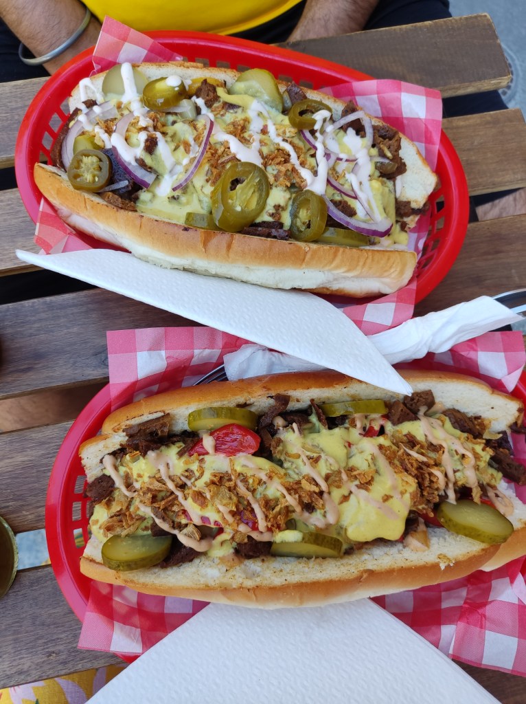 Two big cheesesteak sandwiches, each in a plastic red basket with red and white checked paper. The sandwiches are huge, filled with seitan and cheese. One is topped with fried onions, the other with red onions and jalapenos