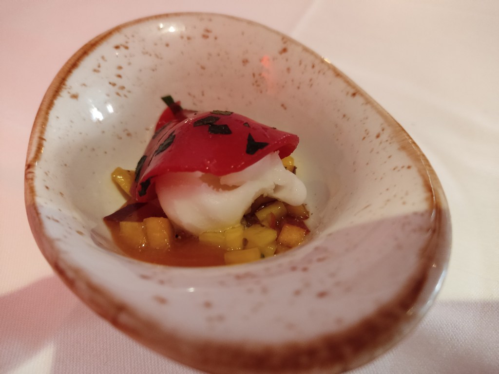 A small dish containing a small scoop of white sorbet sitting in some fruity juice and small pieces of peach. The sorbet is topped with a pink gelatinous looking disc.