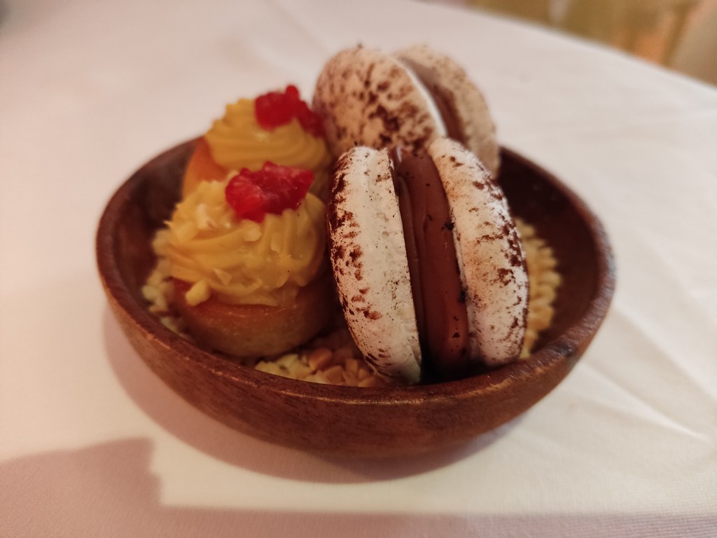 A small wooden dish containing two small cakes topped with yellow cream and a raspberry, and two macarons with white shells and a chocolate cream in the middle.