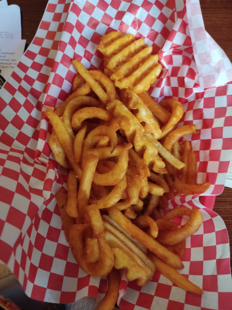 A basket of crispy fries, curly fries, and waffle fries on red and white checked paper