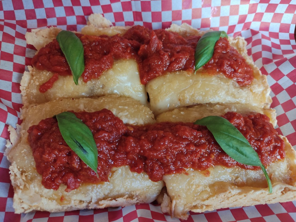 A square pizza cut into quarters covered in vegan cheese and served with tomato sauce scooped on and basil leaves for garnish