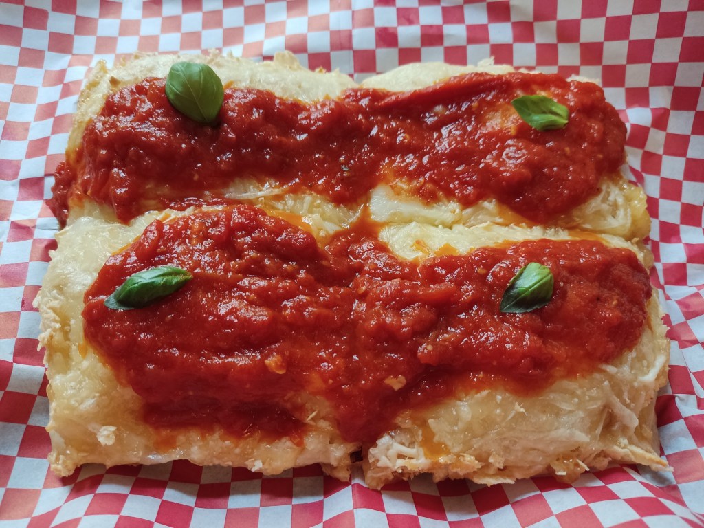 A square pizza cut into quarters covered in vegan cheese and served with tomato sauce scooped on and basil leaves for garnish
