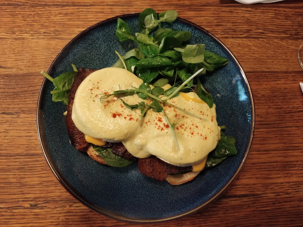 A plate containing two toasted bread buns topped with tofu bacon, tofu eggs, and creamy sauces, with a handful of salad leaves on the side.