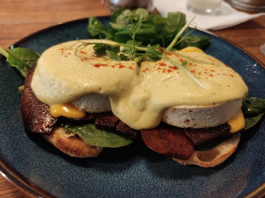 A side view of a plate containing two toasted bread buns topped with tofu bacon, tofu eggs, and creamy sauces, with a handful of salad leaves in the background.
